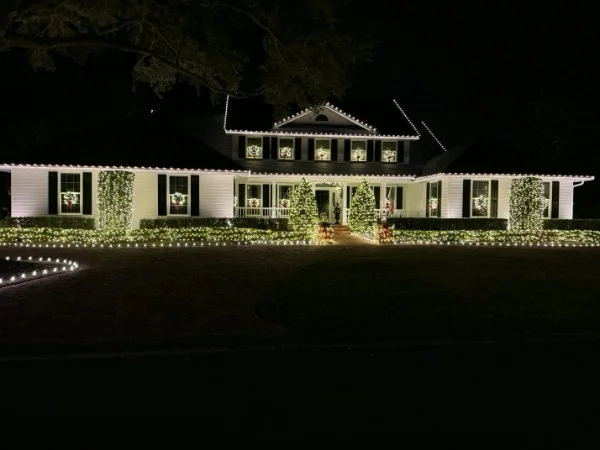 Large white two-story home with white roofline lights, wreaths in the windows, lit hedges, and lit evergreen trees at the entry