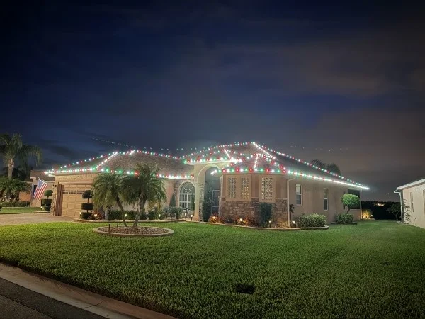 Single-story home at dusk with red, green, and white lights along the roofline, palm trees, and a green lawn