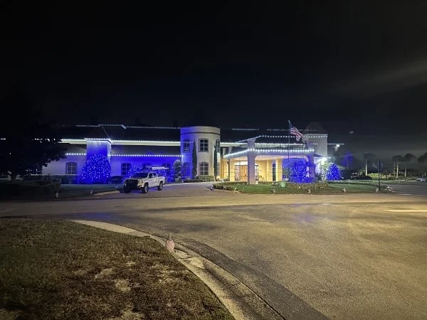 Large clubhouse-style building at night with white roofline lights and blue-lit trees and shrubs beside a circular drive