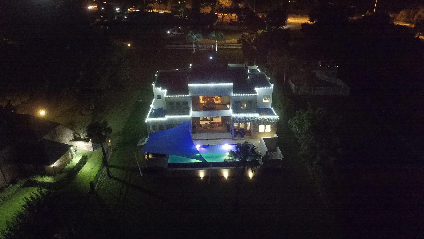 Aerial night view of a large modern home with white lights outlining its rooflines and terraces, a glowing pool, and a blue shade sail
